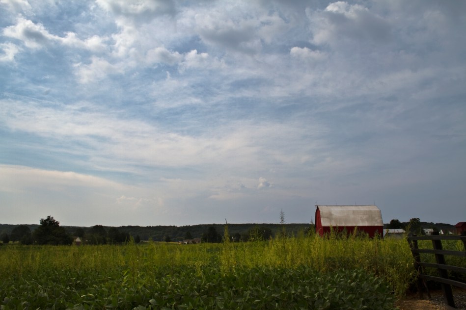 An image of a red barn standing in a field on some rural farmland with dynamic clouds.